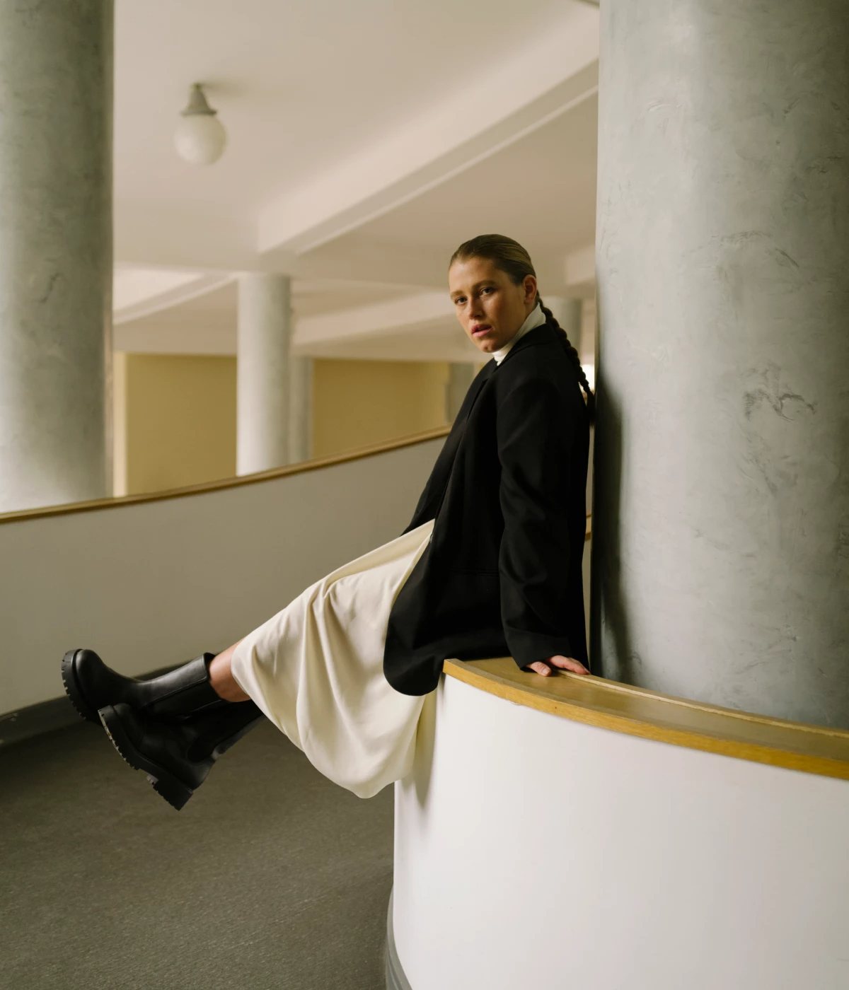 COS — model seated on a curved balustrade, black blazer and ivory skirt with leather boots